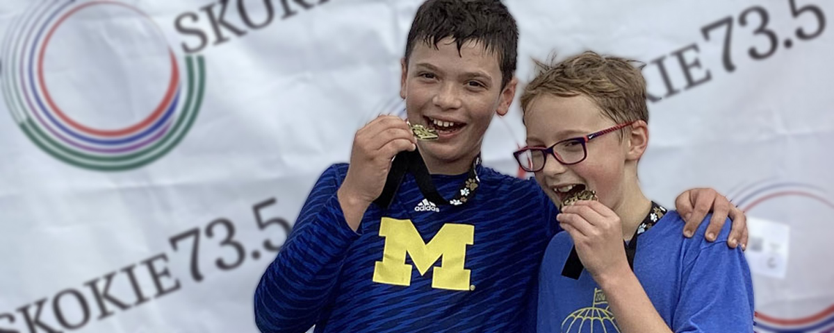 Photo of two 5K race finishers standing in front of a Skokie73.5 backdrop pretending to bite down on their gold medals